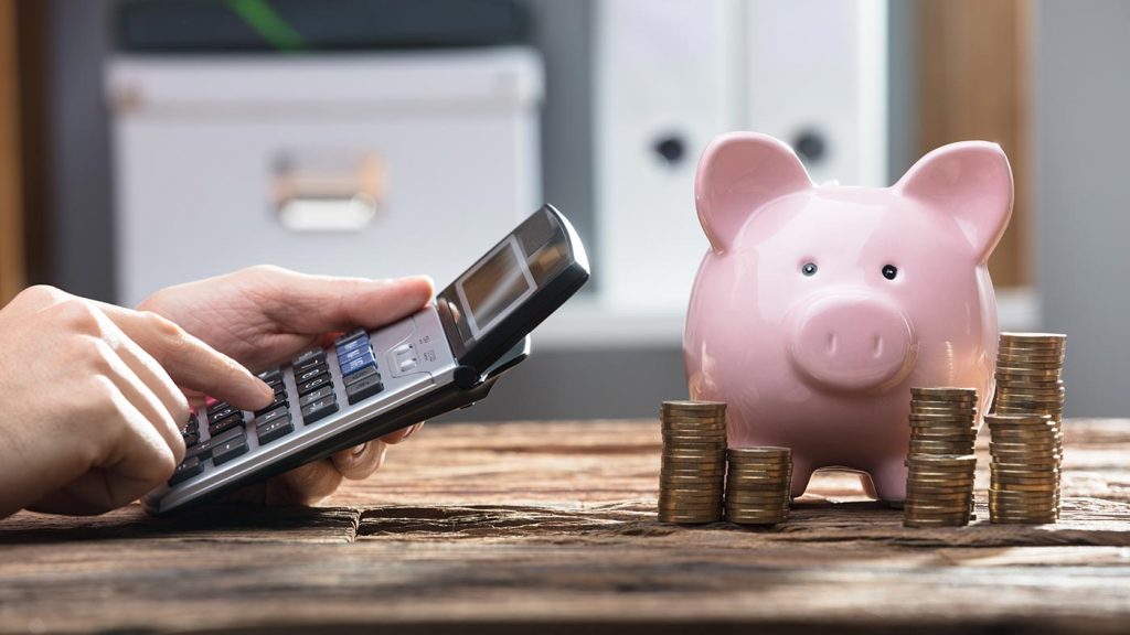 Taschenrechner berechnet das persönliche Budget während auf dem Tisch davor ein rosa Glückschwein und mehrere Münzstapel stehen
